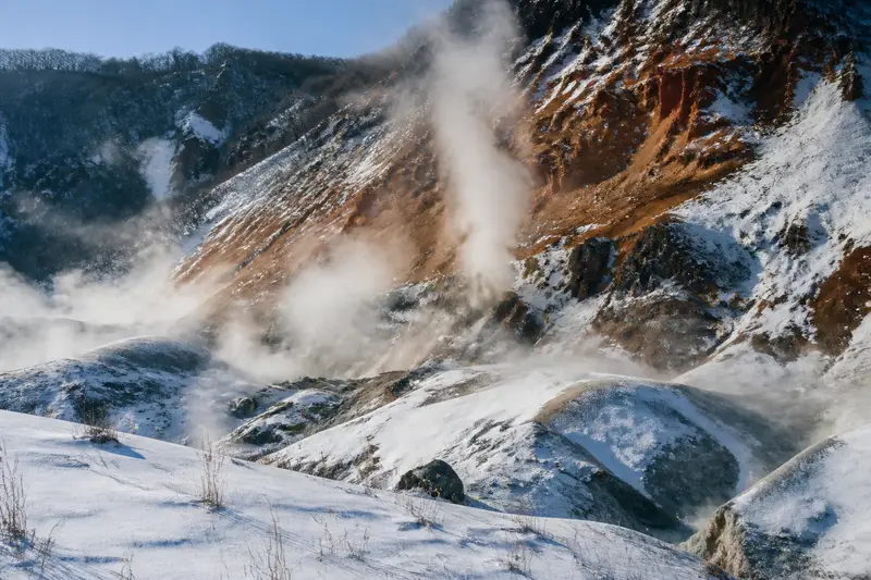 登別地獄谷冬季景象，火山噴氣孔白煙裊裊，雪地與紅褐岩壁