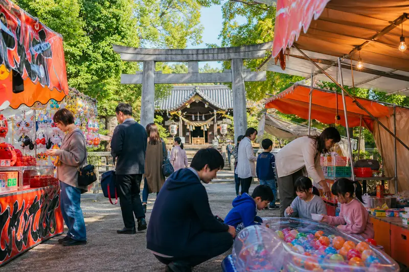 日本白天地方祭典中，親子在神社附近逛攤販與玩遊戲。