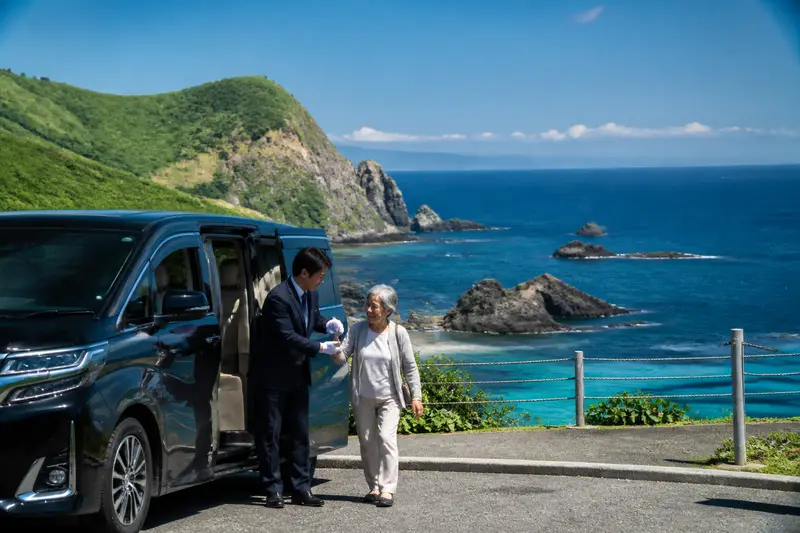 積丹海岸旁的專車，中文司機協助長輩下車看海，背景是積丹藍海灣